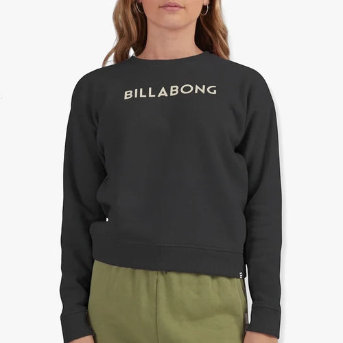 Female model wearing a black Billabong Girls Dancer Crew Sweater with a white logo and olive green sweatpants, viewed from