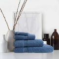 Stack of light blue Colibri Imperial Zerotwist bath sheets on a white surface with decorative vases, branches, and bottles.
