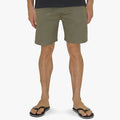 A person wears olive green Dc Boys Rogers shorts and black flip-flops, captured from the waist down against a light