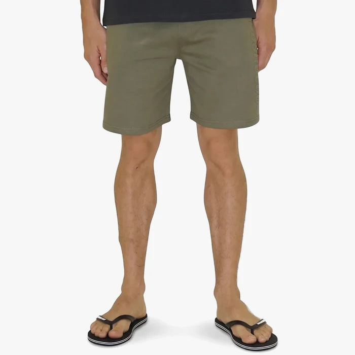 A person wears olive green Dc Boys Rogers shorts and black flip-flops, captured from the waist down against a light